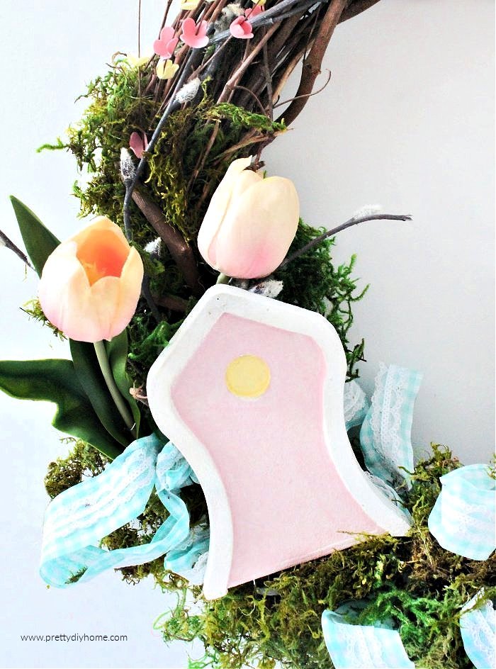 A fairy door attached to a Spring wreath craft as part of a fairy garden.The fairy door is painted a soft pink with white door jam and a yellow winow. It is surrounded by light blue ribbon and tuliips.