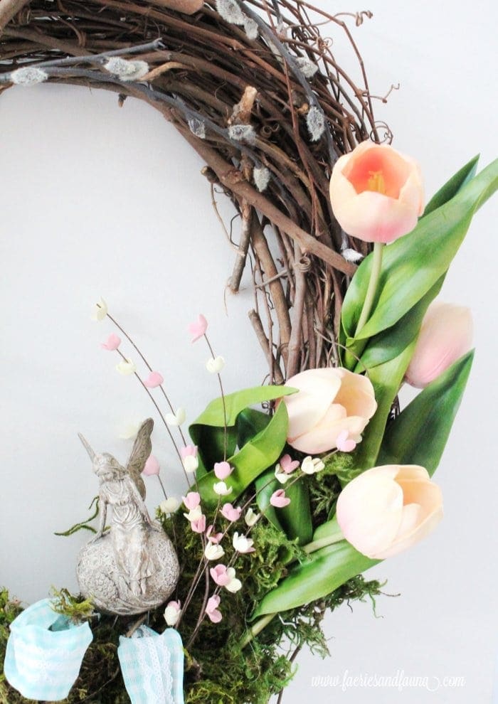 A beautiful clay fairy ornament sitting in the bottom corner of a wreath. The wreath is decorated as a fairy garden with pretty Spring tulips, small pink flowers, soft green moss and a fairy door.