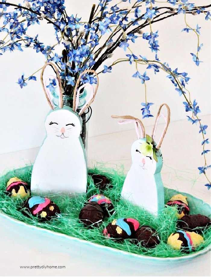 A pair of woodem bunnies sitting on a tray layered with green Easter grass and chocolate eggs.  There is a Easter flower arrangement in the background.