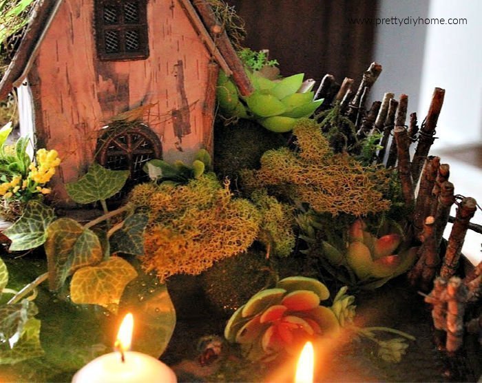 A closeup of a whimsical indoor fairy garden showing faux plants, succulents a fairy garden, fairy twig fence and a water filled pond.