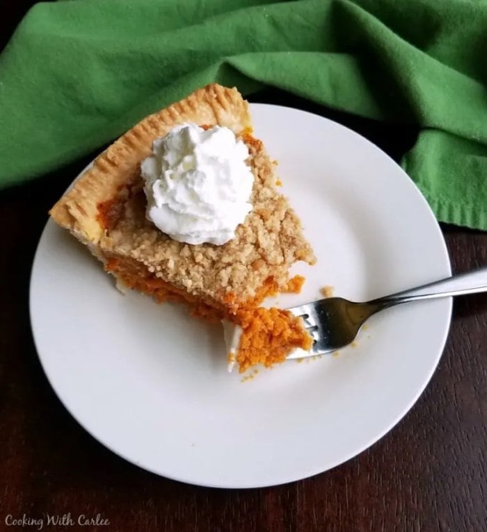 A slice of cinnamon struesel carrot pie on a white plate. The pie has a piece broken off with a fork and there is whipped cream on top.