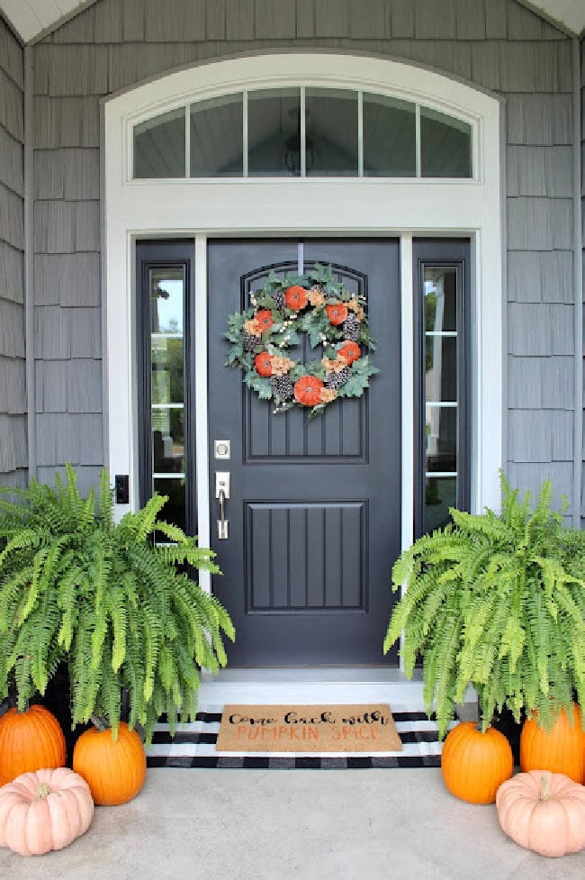 Front porch decorated with fall, the door is black, with two large green fern plants on each side, there and loads of fresh orange pumpkins.