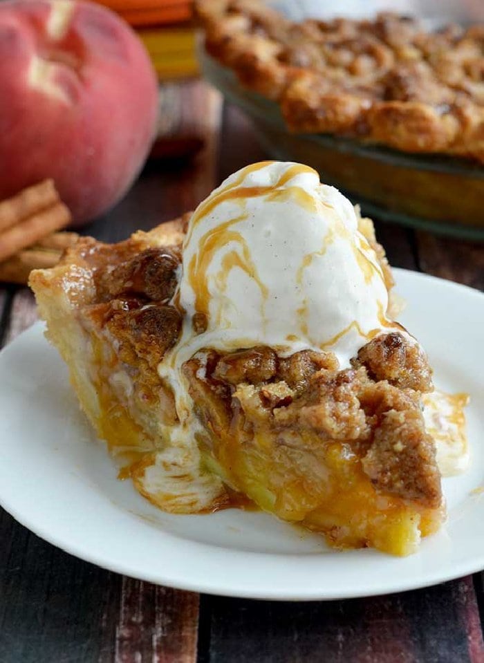 A slice of maple bourbon peach pie covered with ice cream. The peach pie has a crumb top and is being served on a white plate