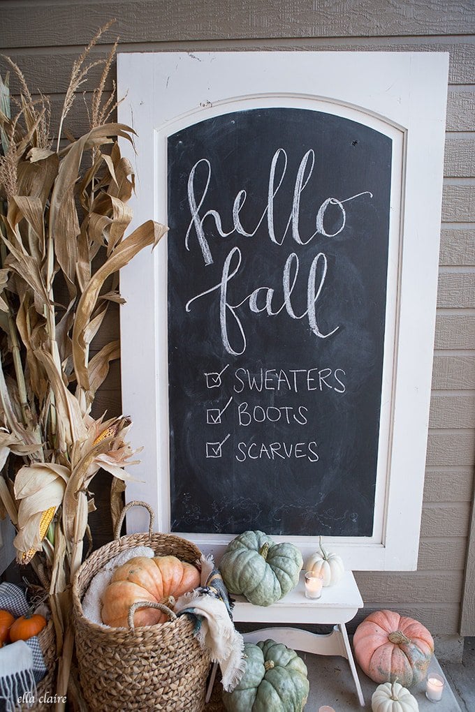 A traditional decorated fall porch with a large chalkboard, baskets, corn stalks, and a basket full of green and orange pumpkins.