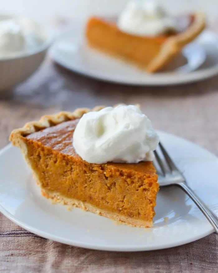 A slice of sweet potato pie served with whipped cream. The pie is sitting on a white plate and their is a fork on the plate as well.