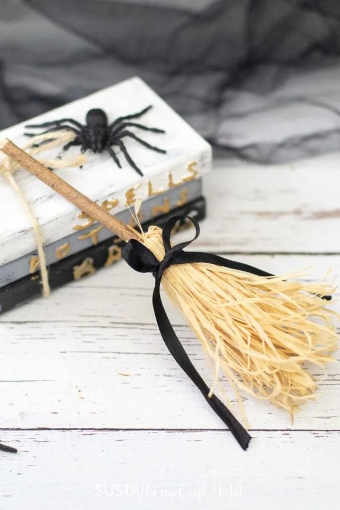 A minature sized witch broom craft made with a small stick and raffia bristles.  The mini witch's broom is tied with a black silk ribbon and is leaning against a stack of black grey and white book stack with a giant spider on top.