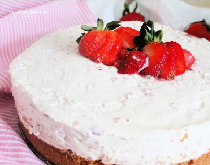 A large whole strawberry rhubarb cheesecake. The no bake cheesecake has a graham cookie crust a thick layer of pink all natural filling, and sliced strawberries on top.