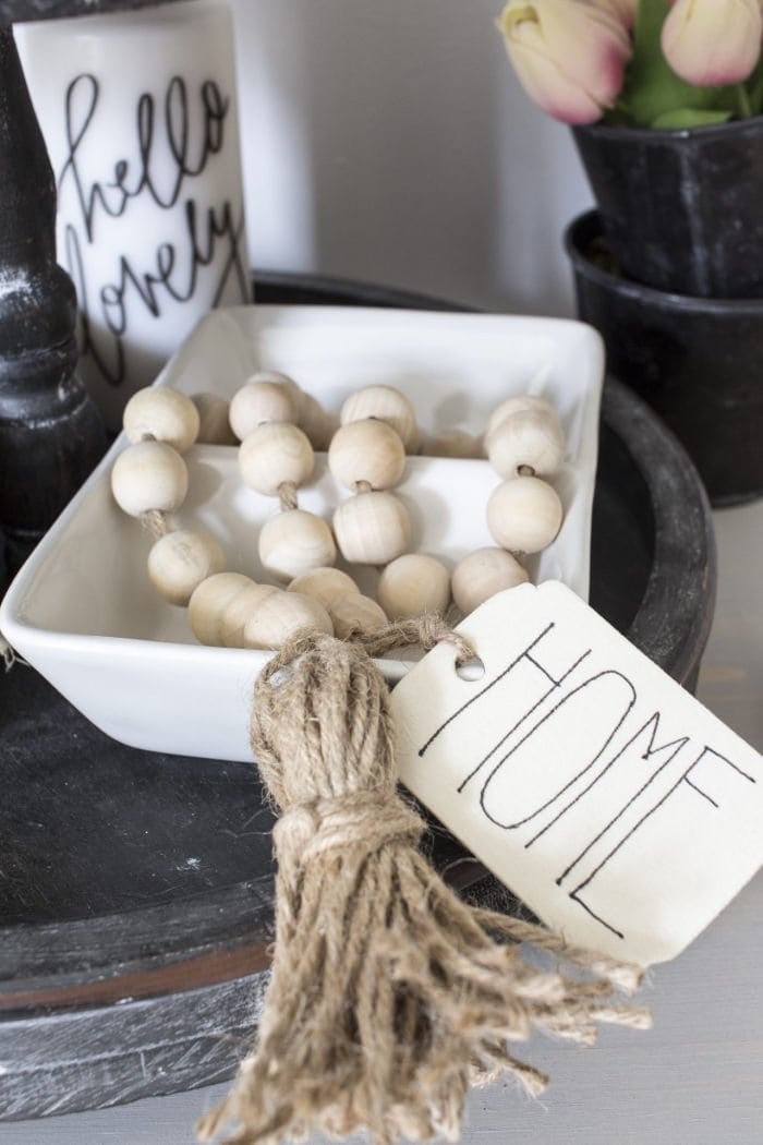 A farmhouse natural wood bead garland with tassles and a label that says home. Showing in a pretty farmhouse display on a grey tray.