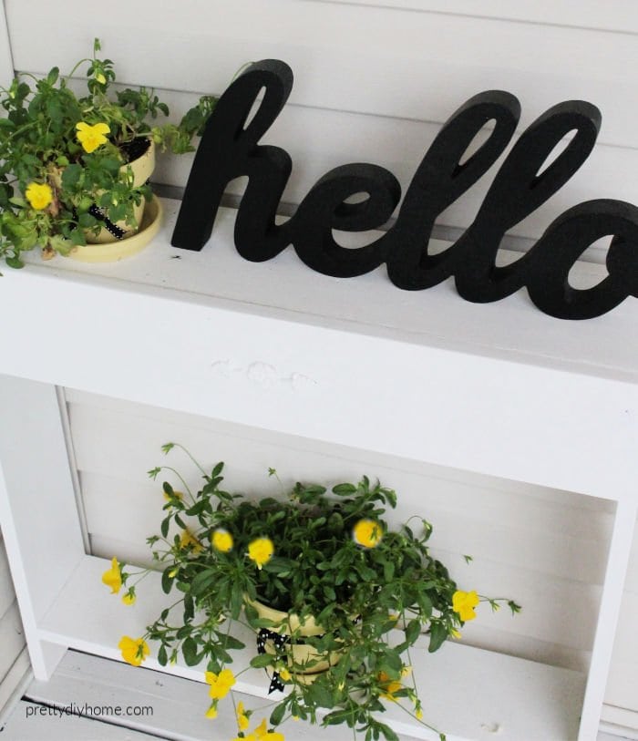 A DIY side table on a front porch with a hello sign in black, and two yellow flower pots filled with pansies.