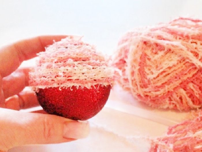 A DIY Christmas ornament ball being held by a hand.  You can see the top of the Christmas tree ornament is covered in pink cotton wool and the bottom half is sparkling red glitter.