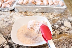 A large white bowl filled with flour and spices, and a coated chicken wing being made using tongs, The baking pans full of chicken wings are in the background.