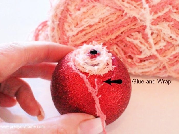 Showing the top of a red dollar tree Christmas ornament thats being wrapped in pink homespun looking cotton wool. The Christmas ball is red and the homespun wool is pink.