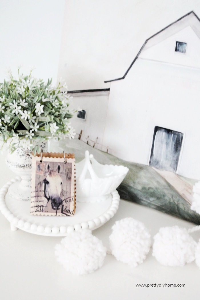 A white DIY wood table riser with white bead edging, holding milk glass, a small flower plant and booklet with a horse cover. Surrounded by a snowball garland and white barn sign backdrop.