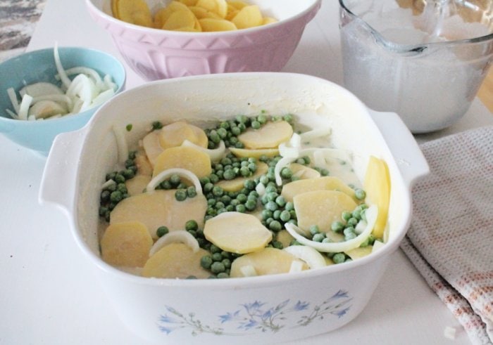 Adding an optional layer of green peas to homemade classic scalloped potato recipe in a large casserole dish with a lid