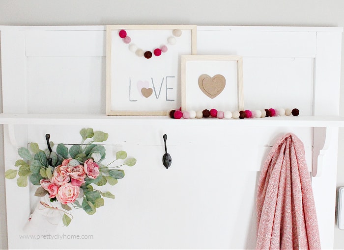 Two wood Valentines with white background, and natural wood frame. One sign says Love the other one has a layered brown Valentine. Both pictures are draped with pretty felt ball garland.