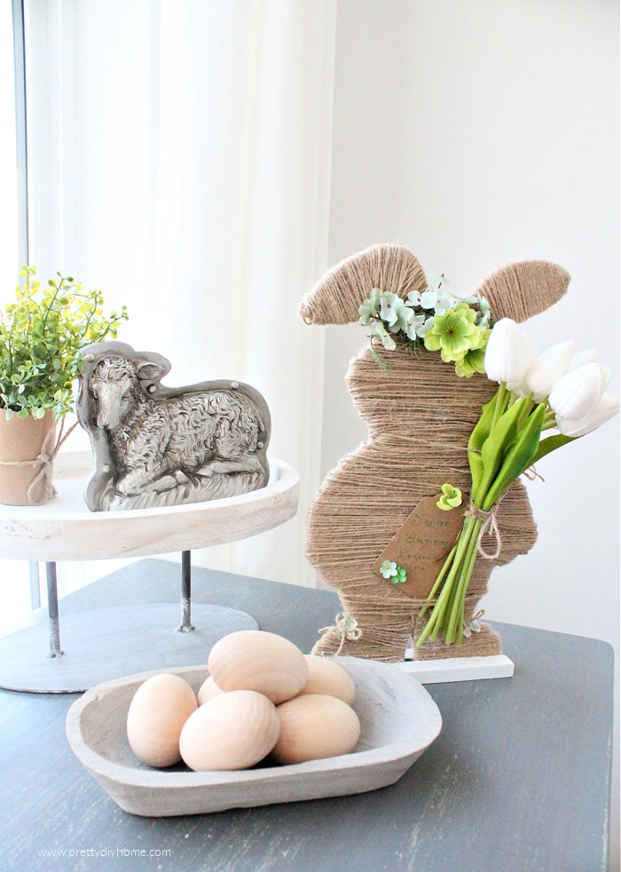 A neutral farmhouse Easter bunny made with twine and faux flowers. He is sitting beside a small bowl full of natural wooden eggs, a vintage sheep chocolate mold and a farmhouse tray.