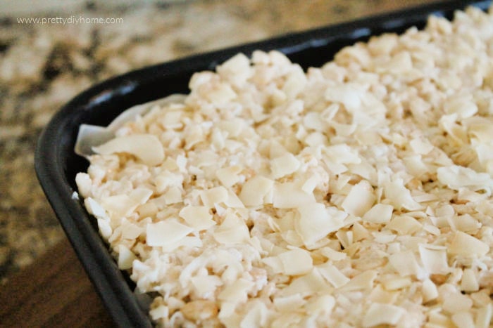 A large tray of layered rice krispie treats with coconut and almonds The squares are cooling in a large black pan and have not been cut yet.