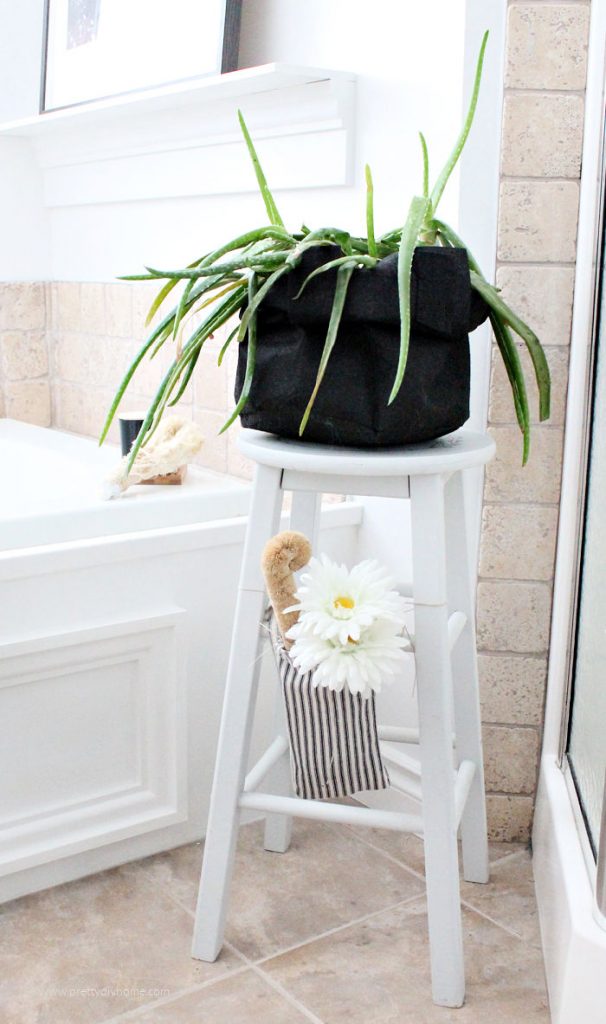 Farmhouse plantstand for the bathroom with an aloe vera plant on top and a scented farmhouse bag with a flower hanging on the side.