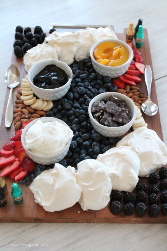 A large wood charcuterie board with homemade mini pavlova, blackberries, strawberries, blueberries, bananas, almonds, peaches and chocolate liquere bottles.