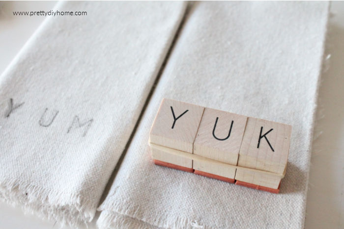 YUK spelled out on with wood stamps. The stamp is bundled together with elastic and is making a DIY stamped fabric napkin craft.