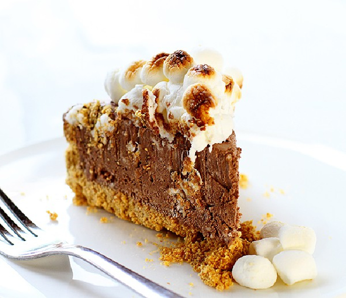 A thick slice of homemade frozen s'mores cheesecake on a white plate with a fork beside it. The cheesecake has a graham crust, thick dense layer of chocolate cheesecake filling, and toasted marshmallow topping.