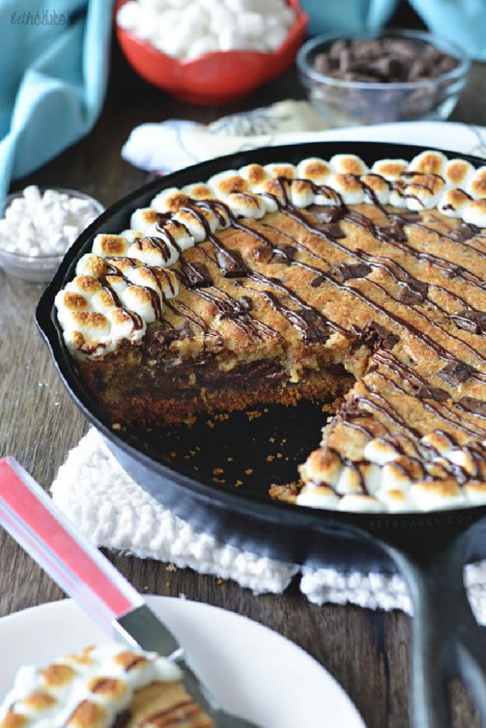 A large caste iron pan sitting on a counter filled with S'more skillet cookie. The large cookie has a slice out of the middle so you can see the layers, and it is edged with toasted mini marshmallows.