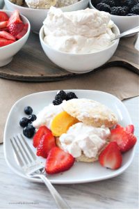 Homemade classic strawberry shortcake served with sweet biscuits, there is a dessert charcuterie board in the background with strawberries, raspberries, peaches, blackberries, blueberries and whipping cream.