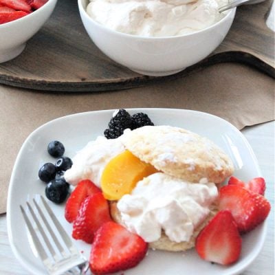 Homemade classic strawberry shortcake served with sweet biscuits, there is a dessert charcuterie board in the background with strawberries, raspberries, peaches, blackberries, blueberries and whipping cream.