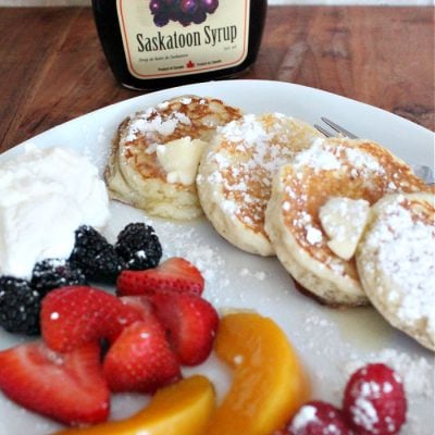 A serving of four silver dollar sized lemon ricotta pancakes dusted with icing sugar and dabs of butter. The pancakes are served on a white plate with an assortment of fresh berries, blueberries, strawberries, blackberries, raspberries and peaches in neat rows.