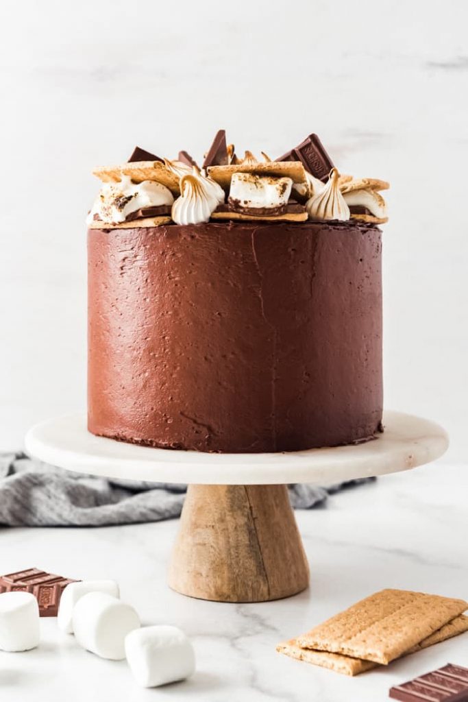 An entire S'mores cake with smooth chocolate icing on a pedestal plate.  The top of the cake is decorated with dollops of marshmallow fluff and big chunks of luscious chocolate.