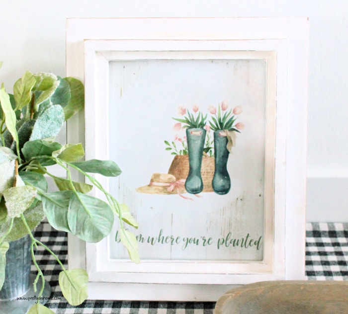 Printable wall art of a Spring garden hat, and rubber boots full of flowers. The printable wall art says bloom where you are planted. There is a pretty green plant sitting beside it and its sitting on farmhouse checked fabric.