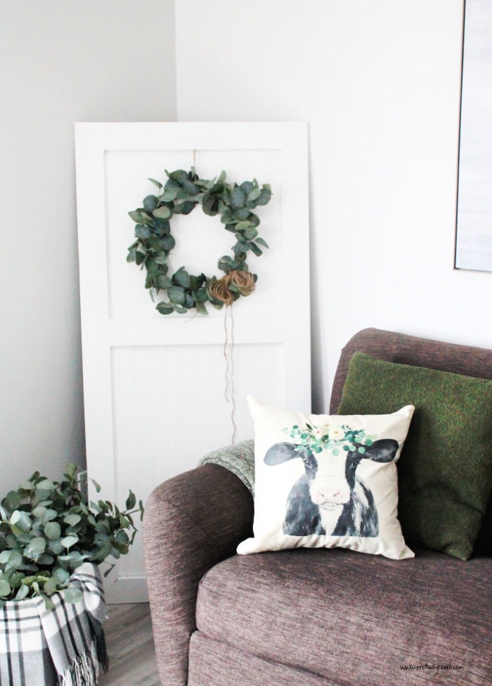 A DIY white door made out of beadboard propped in the corner of a living room. The door has a green eucalyptus wreath hanging from the front. The door is sitting between a dark grey couch and a basket full of branches and a grey plaid throw.