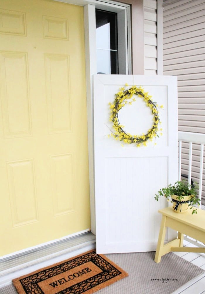 A handmade DIY wooden door made with beadboard, and then painted white. the door decorated with a yellow Spring flower wreath, and is sitting on a Spring front porch with yellow bench beside it.