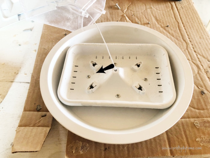 The bottom of a DIY bird feeder idea attached to a dollar store pie plate to make the stand larger. Both pieces are painted white and there is a black arrow emphasizing the placement of the screws.