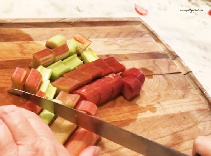 Preparing washed rhubarb for rhubarb recipe. There rhubarb is being cut into 3/4 inch slices on a cutting board.