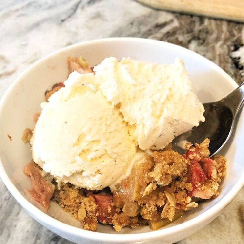 A bowl of rhubarb crisp with ice cream. The rhubarb dessert is in a white bowl with a spoon and you can see the corner of the rhubarb crisp in the back corner.