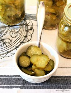 A bowl full of bread and butter pickles surrounded by jars of homemade bread and butter pickles in cans.