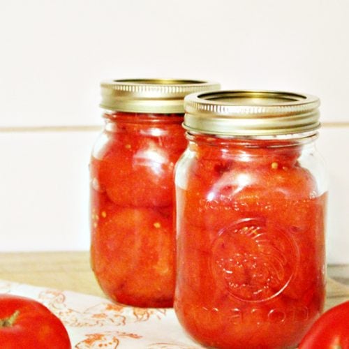 Two jars of canned tomatoes sitting on a countertop with fresh whole tomatoes.