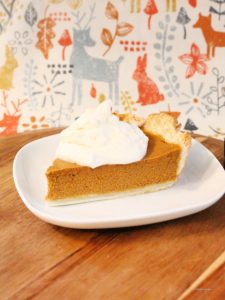 A large slice of pumpkin pie recipe from scratch on a white plate. The slice of pie has whipping cream on top and a pretty fall themed tea towel background.