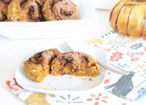 A plate with a fresh pumpkin scone cut open so you can see the cinnamon swirl center. There is a bowl of pumpkin scones in the background and a pretty orange glass pumpkin.