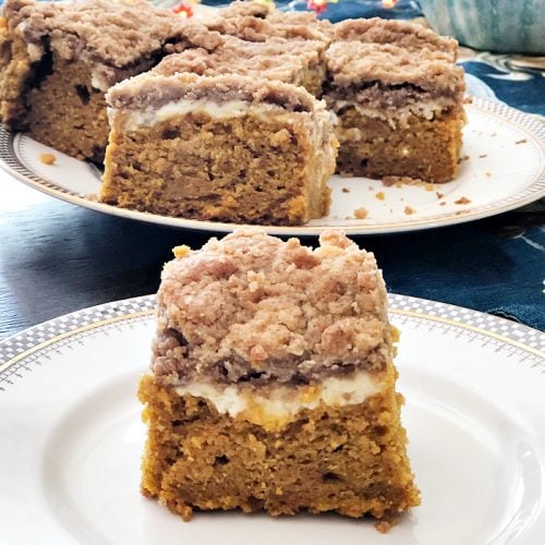 A plate with a thick slice of pumpkin cake. The cake has three layers, orange pumpkin cake bottom, then a light cream cheese center, finally golden brown struesel topping. There is a large plate of the pumpkin cake in the background.