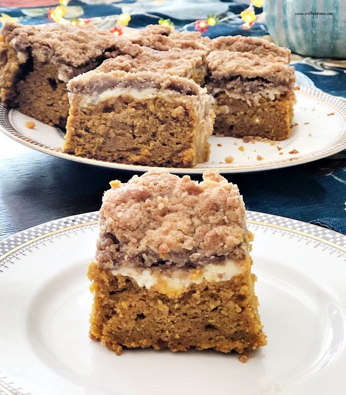 A plate with a thick slice of pumpkin cake. The cake has three layers, orange pumpkin cake bottom, then a light cream cheese center, finally golden brown struesel topping. There is a large plate of the pumpkin cake in the background.