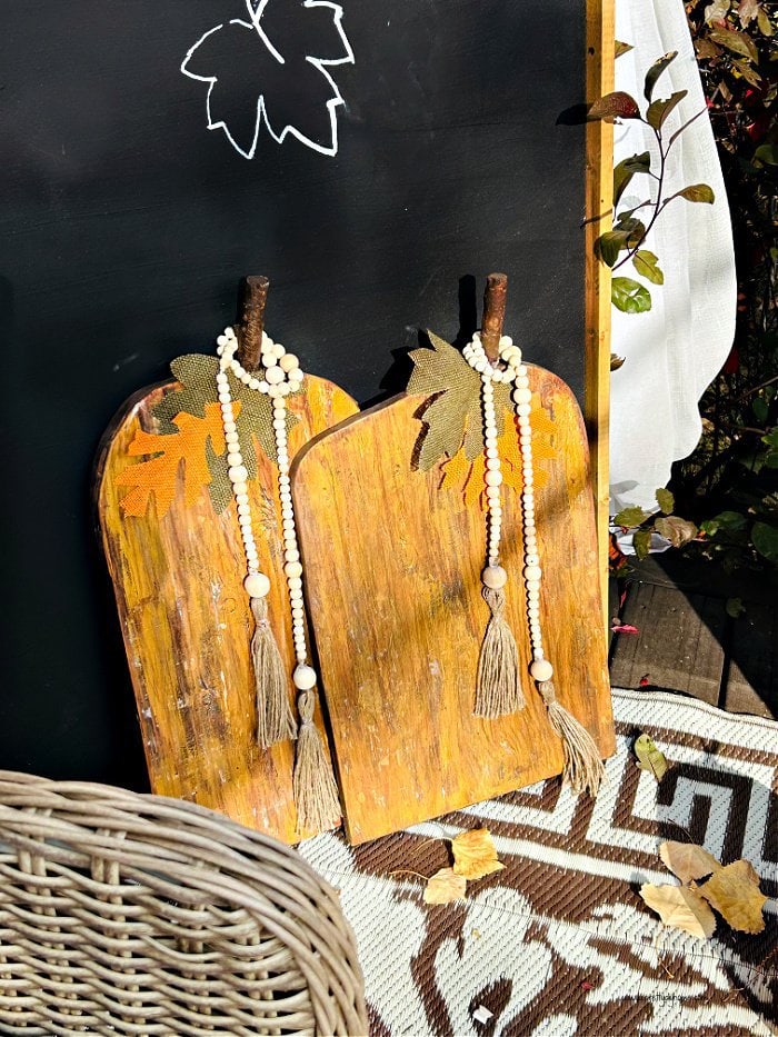 Two Wooden pumpkins in gold and brown with green burlap leaves, and farmhouse bead garlands.