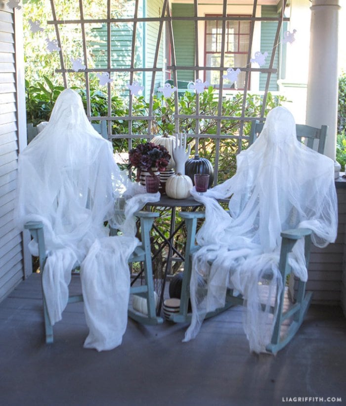 Two Halloween outdoor decorations large size ghostss casually sitting on the end of a porch in rocking chairs.