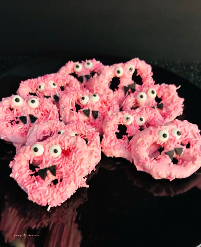 Easy pretzel for Halloween idea with pink candy coating, licorice fangs and white eyeballs. The bright pink Halloween treats are sitting on a black plate.