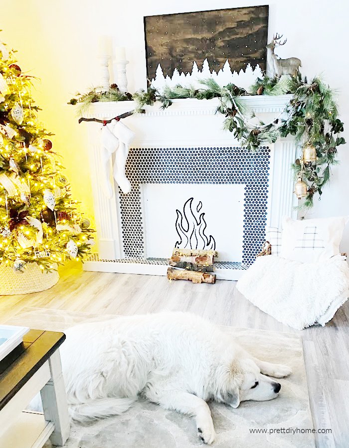 A Christmas decor for the mantel with cushions, greenery, gold bells, twinkling lights, a sign and candles all in neutral white, green, and brown. There is a large puppy relaxing in front of the fireplace.