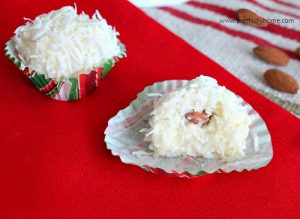 Two coconut snowballs one is whole the other has a bite out of it showing a almond center. The coconut snowballs are sitting on a red fabric background.