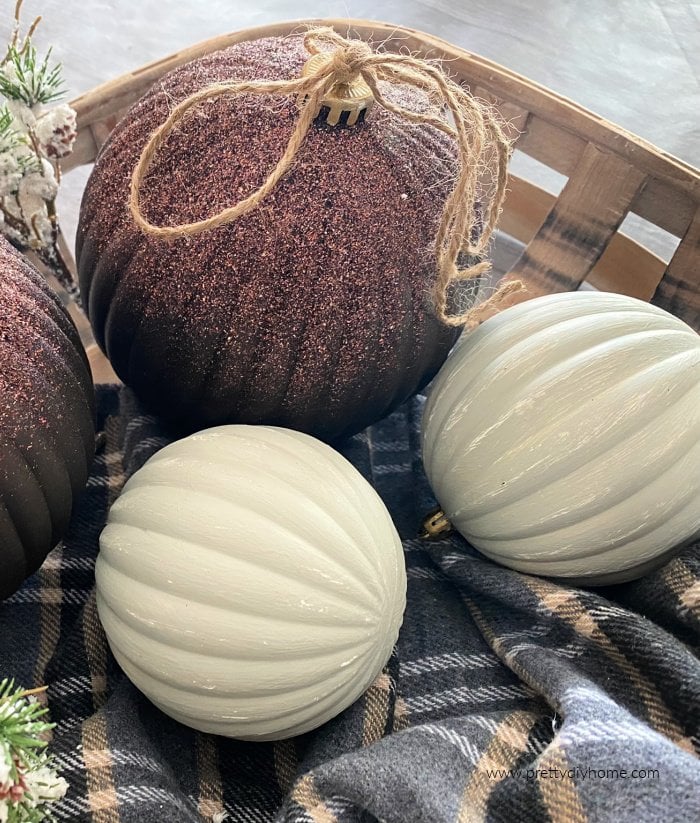 Two giant Christmas ornaments in chocolate brown paint and glitter with twine bows. The two brown Christmas ornaments are sitting in a basket with two soft green painted DIY Christmas ornaments that have been distressed for a soft cottage or farmhouse look.