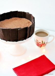 A thick no bake chocolate cheesecake sitting on a white pedestal cake plate. The chocolate cheesecake has a chocolate collar encircling it.