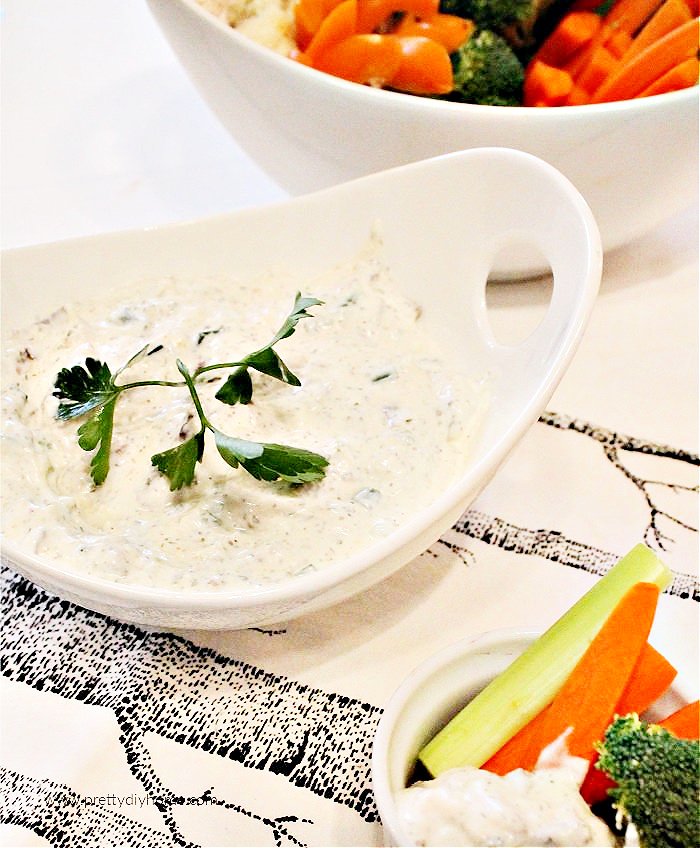 A large bowl filled with homemade ranch dip, there is a small bowl with fresh cut up vegetables beside it and a large serving dish full of fresh vegetables like carrots, brocolli and celery behind the dip bowl.